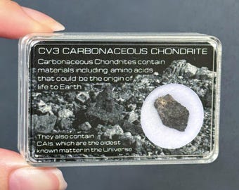 May include: A clear rectangular plastic display case containing a small, dark, textured CV3 carbonaceous chondrite meteorite fragment. The label features text about amino acids and CAIs, with a black and white image of rocky terrain.