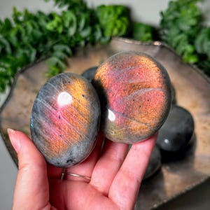 Pode incluir: Duas pedras de labradorite polidas de formato oval com uma cor base cinzento-escura e reflexos iridescentes vibrantes em laranja, rosa e dourado. As pedras têm uma superfície lisa e refletora que capta a luz, revelando camadas minerais internas.
