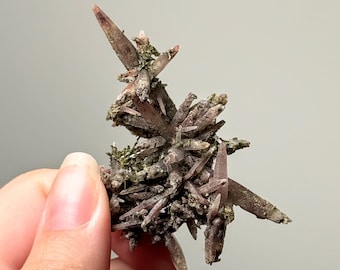 May include: A cluster of pointed, dusty mauve and brown mineral crystals with textured, mossy green inclusions. The formation features multiple sharp, elongated crystal points radiating from a central, rugged base with earthy, muted tones.