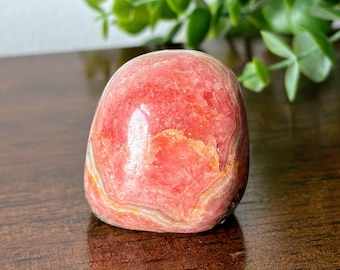 May include: A polished, rounded stone featuring a vibrant mix of coral, salmon, and soft pink hues. The surface displays intricate, layered banding patterns with translucent crystalline sections and subtle white mineral veins throughout the structure.