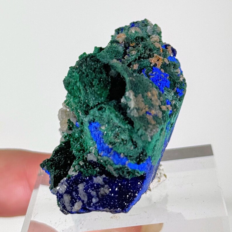 May include: A raw mineral specimen featuring a combination of deep forest green malachite and vibrant royal blue azurite crystals. The cluster displays a textured, crystalline surface with small, light-colored mineral inclusions throughout.
