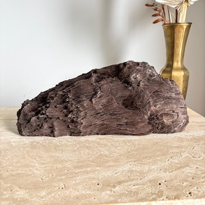 May include: A dark brown, textured rock formation with a rough, uneven surface. The rock is sitting on a light beige surface.