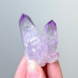 May include: A cluster of three translucent amethyst crystal points with a natural jagged texture. The crystals exhibit a gradient of purple hues, transitioning from deep violet at the sharp, pointed tips to a lighter, clearer base.
