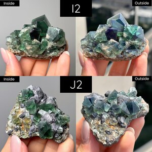 May include: Two clusters of raw fluorite crystals labeled I2 and J2, shown in both indoor and outdoor lighting. The specimens feature translucent green and deep purple cubic crystals embedded in a rough, greyish-brown mineral matrix base.