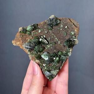 May include: A natural mineral specimen featuring clusters of dark green crystals on a brown matrix. The crystals have a glassy luster and are irregularly shaped. The matrix has a rough, textured surface. The overall appearance is earthy and organic.