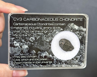 May include: A rectangular clear plastic display case containing a small, dark, textured meteorite fragment centred on a white circular foam insert. The case features printed text: CV3 CARBONACEOUS CHONDRITE, describing amino acids and CAIs.