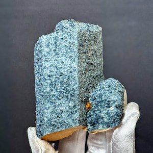 May include: A textured, blue-grey mineral specimen with a rectangular shape and a smaller, attached piece. The mineral has a rough surface and a small, orange-brown inclusion. The specimen is held by a gloved hand against a dark background.