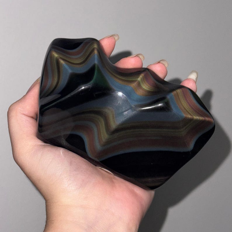 May include: A polished, irregular-shaped piece of rainbow obsidian featuring a smooth, glossy black surface with distinct, layered bands of iridescent colors including gold, blue, and green that create a wavy, rippled pattern across the stone.