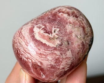 May include: A polished rhodochrosite stone featuring a smooth, rounded shape with a vibrant mix of deep rose, dusty pink, and creamy white swirling patterns. The surface displays natural crystalline textures and intricate, layered mineral bands.