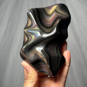 May include: A polished, irregular-shaped stone featuring a deep black base color with swirling, iridescent bands of gold, green, and purple. The surface is smooth and reflective, showcasing a fluid, wave-like pattern throughout the entire object.