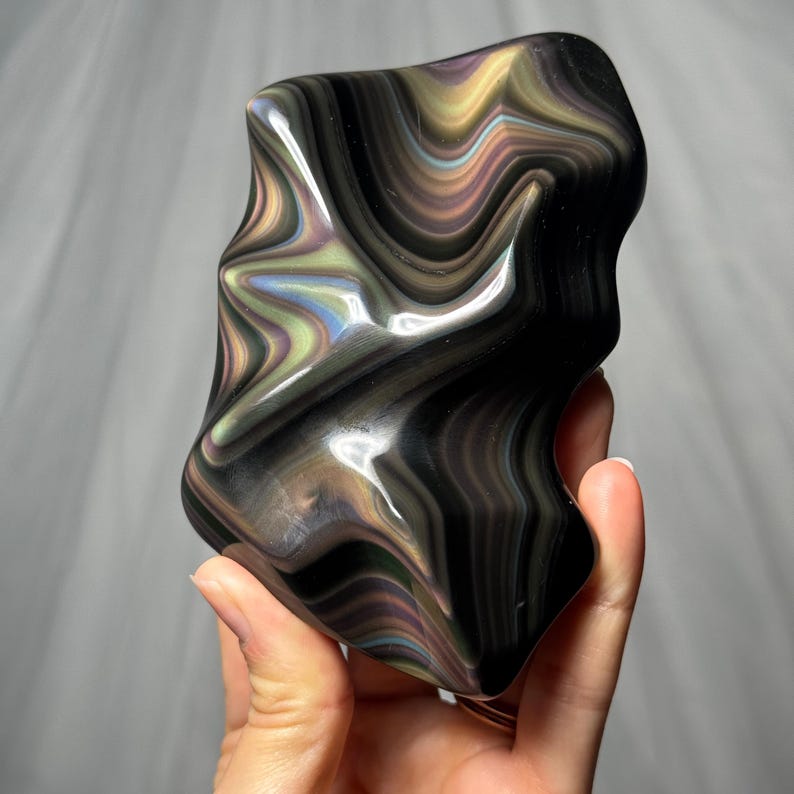 May include: A polished, irregular-shaped stone featuring a deep black base color with swirling, iridescent bands of gold, green, and purple. The surface is smooth and reflective, showcasing a fluid, wave-like pattern throughout the entire object.