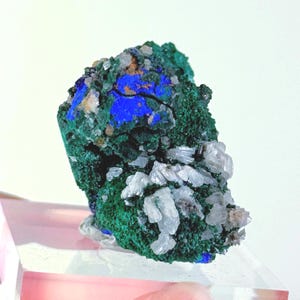 May include: A raw mineral specimen featuring a cluster of deep green malachite, vibrant blue azurite patches, and small, translucent white crystalline formations. The rock has a textured, uneven surface with distinct color variations throughout.