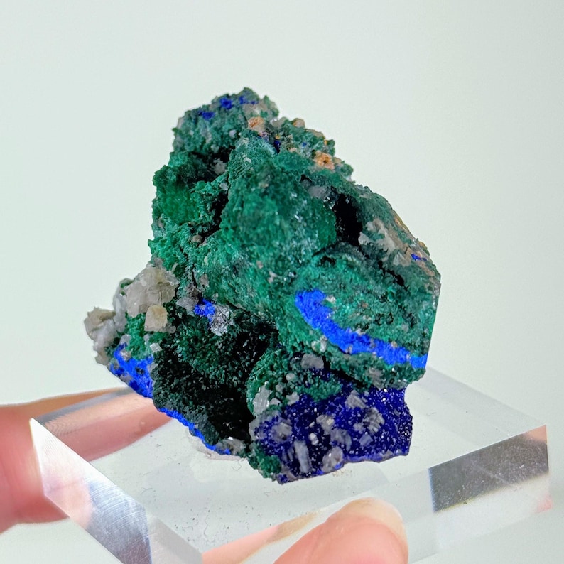 May include: A raw mineral specimen featuring a cluster of deep green malachite and vibrant blue azurite crystals. The geological formation displays a rough, textured surface with contrasting color zones and small, sparkling crystalline inclusions.