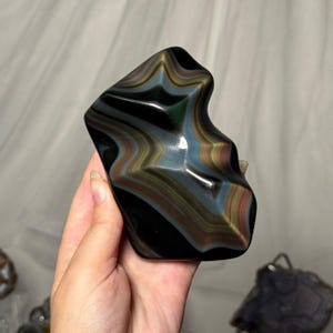 May include: A polished, irregularly shaped obsidian stone featuring a smooth, reflective surface with distinct, layered bands of iridescent rainbow colors including gold, green, and blue hues that create a wavy, multi-dimensional visual effect.