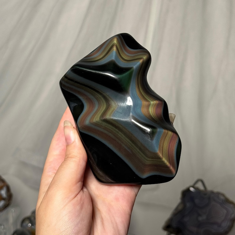 May include: A polished, irregularly shaped obsidian stone featuring a smooth, reflective surface with distinct, layered bands of iridescent rainbow colors including gold, green, and blue hues that create a wavy, multi-dimensional visual effect.