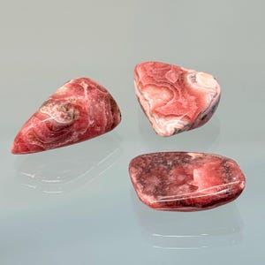 May include: Three polished rhodochrosite stones in shades of pink, red, and white. The stones have a smooth, polished surface and a variety of shapes, including a triangular, rounded, and rectangular form. The stones are displayed on a reflective surface.