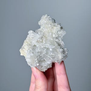 Könnte beinhalten: Ein Cluster aus klaren, kristallinen Mineralformationen. Die Kristalle sind hauptsächlich rechteckig und gruppiert, wodurch eine strukturierte, hellfarbige Gesteinsformation entsteht. Das Mineral wird in einer Hand vor grauem Hintergrund gehalten.