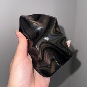 May include: A polished, irregularly shaped stone featuring a smooth, glossy surface with swirling bands of deep black, dark brown, and subtle translucent gray tones that create a fluid, layered pattern throughout the entire object.