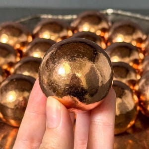 Puede incluir: Un objeto esférico único con un acabado metálico liso y reflectante de color cobre. La superficie está altamente pulida, creando reflejos brillantes y sombras profundas que enfatizan su forma redonda y su textura metálica.