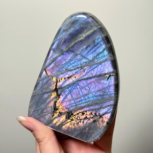 Puede incluir: Una piedra de labradorita pulida, con una parte superior redondeada y una base plana. La piedra muestra un vibrante juego de colores, con tonos de púrpura, azul y dorado. La superficie tiene vetas naturales y un acabado liso y brillante.