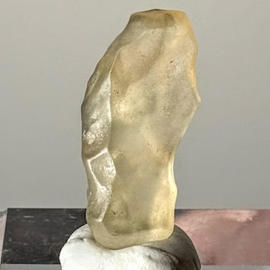 Puede incluir: Una escultura abstracta translúcida de color beige, con una superficie rugosa y texturizada. La escultura tiene una forma irregular y está montada sobre una base blanca. El fondo es gris neutro.