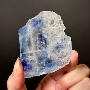May include: A translucent, crystalline mineral with streaks of deep blue and white. The rough-edged, rectangular shape of the mineral is held in a hand against a dark background. The mineral has a layered appearance.