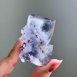 Puede incluir: Un espécimen de fluorita cristalina translúcida. El mineral presenta una estructura en capas con tonos de púrpura y blanco, acentuados por pequeñas inclusiones de color marrón rojizo. Los bordes del cristal son ásperos e irregulares, con un aspecto ligeramente esmerilado.