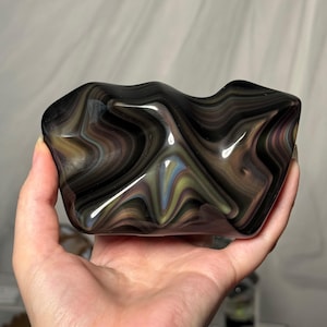 May include: A smooth, polished glass sculpture featuring a swirling, marbled pattern of deep black, charcoal gray, and subtle iridescent highlights of muted green and purple, with an irregular, organic shape and a high-gloss, reflective finish.