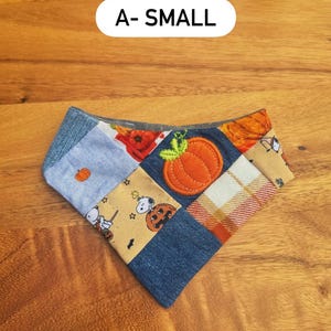 May include: A small triangular pet bandana featuring a patchwork design of denim, plaid, and tan fabric with Snoopy and pumpkin prints. An orange fabric pumpkin appliqu&eacute; is centered on the fabric. The text A- SMALL is visible above.