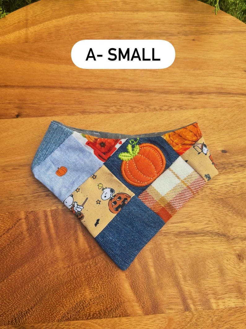 May include: A small triangular pet bandana featuring a patchwork design of denim, plaid, and tan fabric with Snoopy and pumpkin prints. An orange fabric pumpkin appliqu&eacute; is centered on the fabric. The text A- SMALL is visible above.