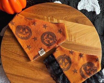 Op de afbeelding: Twee oranje huisdierbandana's met een donkerbruin patroon van pompoengezichten en sterren. Eén bandana heeft een eenvoudige driehoekige rand, terwijl de andere is afgewerkt met een zwarte kanten ruche aan de onderkant. Beide items hebben een label.