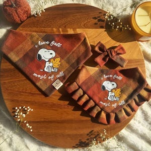 May include: Two plaid pet bandanas in orange, brown, and yellow featuring a Snoopy graphic holding a leaf with the text "i love fall most of all," accompanied by a matching plaid bow accessory. The fabric has a warm, autumnal color palette.