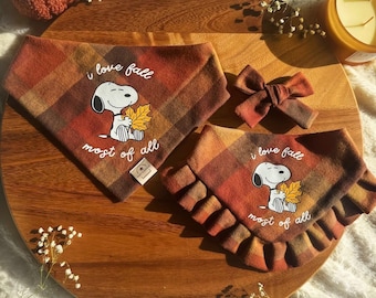 May include: Two plaid pet bandanas in orange, brown, and yellow featuring a Snoopy graphic holding a leaf with the text "i love fall most of all," accompanied by a matching plaid bow accessory. The fabric has a warm, autumnal color palette.