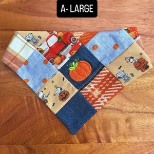 Puede incluir: Un pa&ntilde;uelo triangular para mascotas con un dise&ntilde;o de patchwork de telas con motivos oto&ntilde;ales, que incluye un cami&oacute;n rojo, calabazas y personajes de Snoopy. El texto A-LARGE est&aacute; impreso en blanco sobre un fondo negro arriba.
