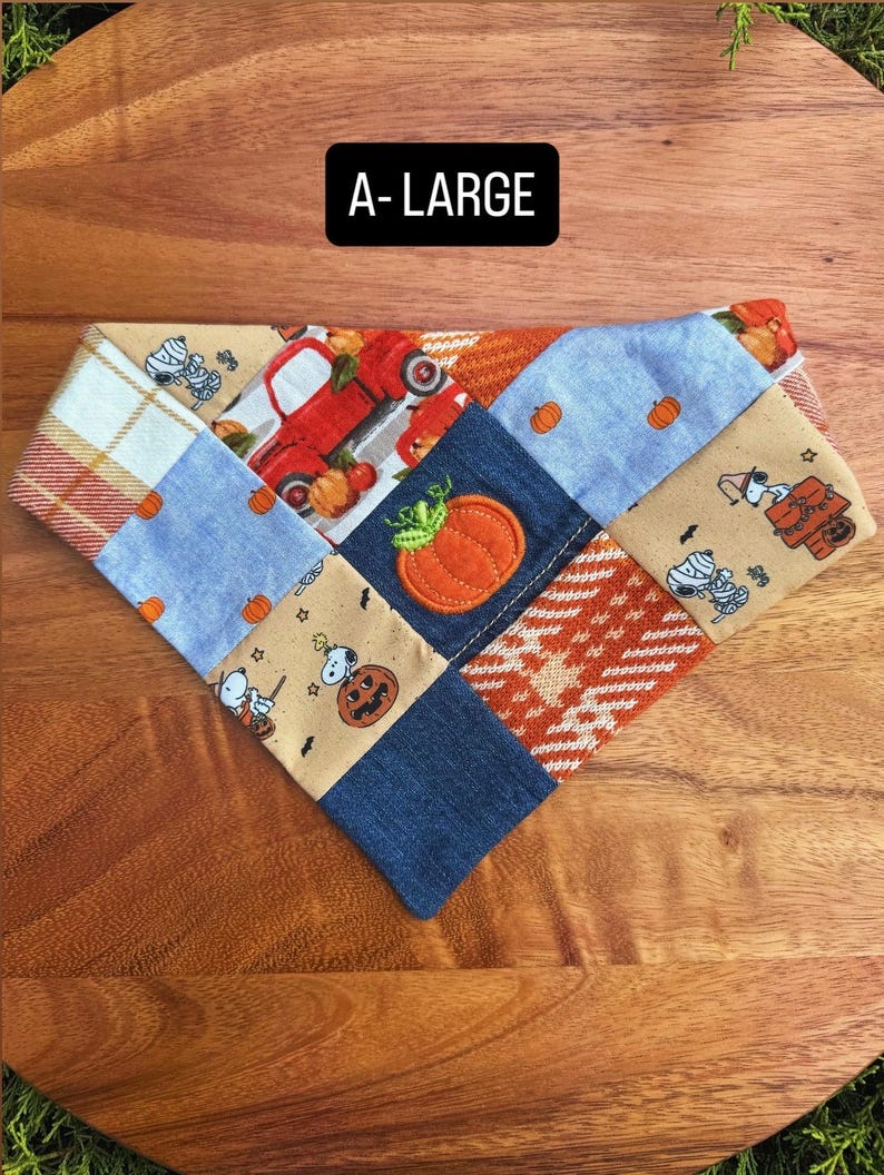 Puede incluir: Un pa&ntilde;uelo triangular para mascotas con un dise&ntilde;o de patchwork de telas con motivos oto&ntilde;ales, que incluye un cami&oacute;n rojo, calabazas y personajes de Snoopy. El texto A-LARGE est&aacute; impreso en blanco sobre un fondo negro arriba.