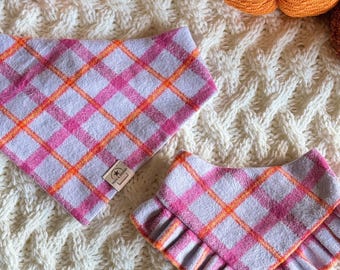 Op de afbeelding: Twee hondenbandana's met een roze, oranje en wit geruit patroon. Eén is driehoekig, de andere heeft een gerimpelde rand. Ze liggen op een witte kabelgebreide deken. Een pompoen en een kaars staan op de achtergrond.