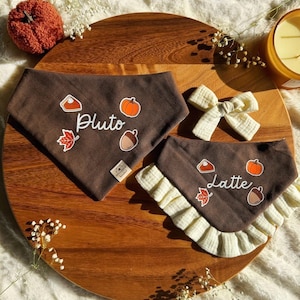 May include: Two brown fabric pet bandanas featuring white text reading Pluto and Latte, decorated with pumpkin, pie, leaf, and acorn graphics. One bandana has a ruffled cream-colored trim, and a small cream-colored fabric bow is included in the set.