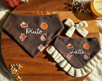 May include: Two brown fabric pet bandanas featuring white text reading Pluto and Latte, decorated with pumpkin, pie, leaf, and acorn graphics. One bandana has a ruffled cream-colored trim, and a small cream-colored fabric bow is included in the set.