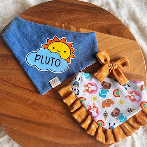 May include: A blue denim pet bandana featuring a yellow sun and a light blue cloud with the text PLUTO, alongside a white pet bandana with a ruffled mustard yellow border, a pattern of dogs, rainbows, and ice cream, and a matching mustard yellow bow.