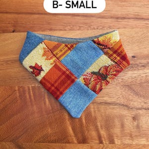 May include: A triangular pet bandana featuring a patchwork design of blue denim fabric, orange and red plaid fabric, and a woven autumn leaf pattern. The text B- SMALL is printed in black on a white rounded rectangular background above it.