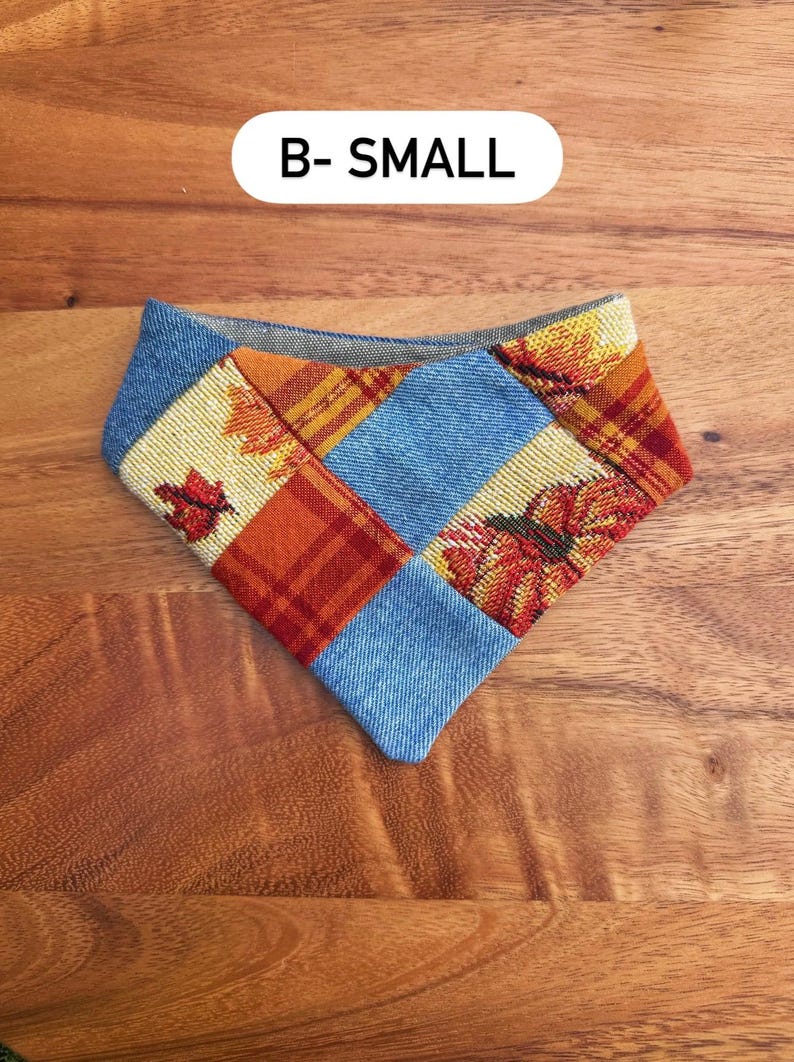 May include: A triangular pet bandana featuring a patchwork design of blue denim fabric, orange and red plaid fabric, and a woven autumn leaf pattern. The text B- SMALL is printed in black on a white rounded rectangular background above it.