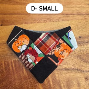 May include: A triangular pet bandana featuring a patchwork design with black fabric, orange pumpkin illustrations, a red and brown plaid pattern, and a cartoon lion character. The text D- SMALL is printed in black on a white background above the item.