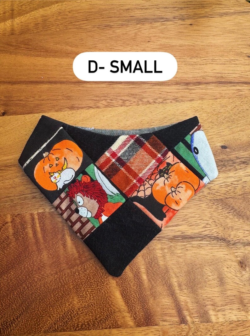 May include: A triangular pet bandana featuring a patchwork design with black fabric, orange pumpkin illustrations, a red and brown plaid pattern, and a cartoon lion character. The text D- SMALL is printed in black on a white background above the item.