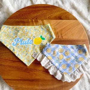 May include: Two triangular pet bandanas. One features a yellow and blue floral pattern with the name Pluto in blue script and a yellow lemon graphic. The second bandana has a blue and white gingham pattern with lemon slices and white lace trim.