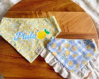 May include: Two triangular pet bandanas. One features a yellow and blue floral pattern with the name Pluto in blue script and a yellow lemon graphic. The second bandana has a blue and white gingham pattern with lemon slices and white lace trim.