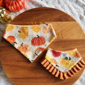 May include: Two dog bandanas with a pumpkin pattern on a wooden surface. The bandanas are white with orange, yellow, and red pumpkins. One bandana is triangular, and the other has an orange ruffle.