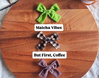 Op de afbeelding: Drie stoffen haarstrikken verticaal gerangschikt. De bovenste strik is limoengroen met witte stippen en het label Matcha Vibes. De middelste strik is zwart en beige geblokt met het label But First, Coffee. De onderste strik is rood en zwart geruit met het label Boo's and Brews.