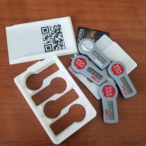 May include: A white plastic holder with three slots for holding key-shaped RSA SecurID tokens. The tokens are gray with red buttons and display a six-digit code. The holder is on a wooden surface.