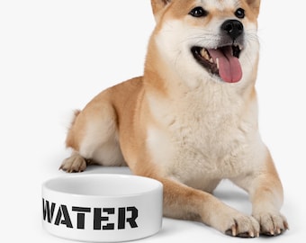 Puede incluir: Un cuenco de cerámica blanco para perros con la palabra "WATER" impresa en letras negras. Un perro Shiba Inu está sentado junto al cuenco.