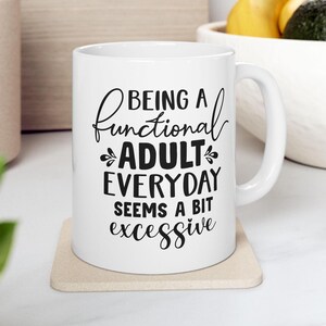 Może przedstawiać: Biały ceramiczny kubek do kawy z czarnym nadrukiem, na którym widnieje napis "Being a functional adult everyday seems a bit excessive".